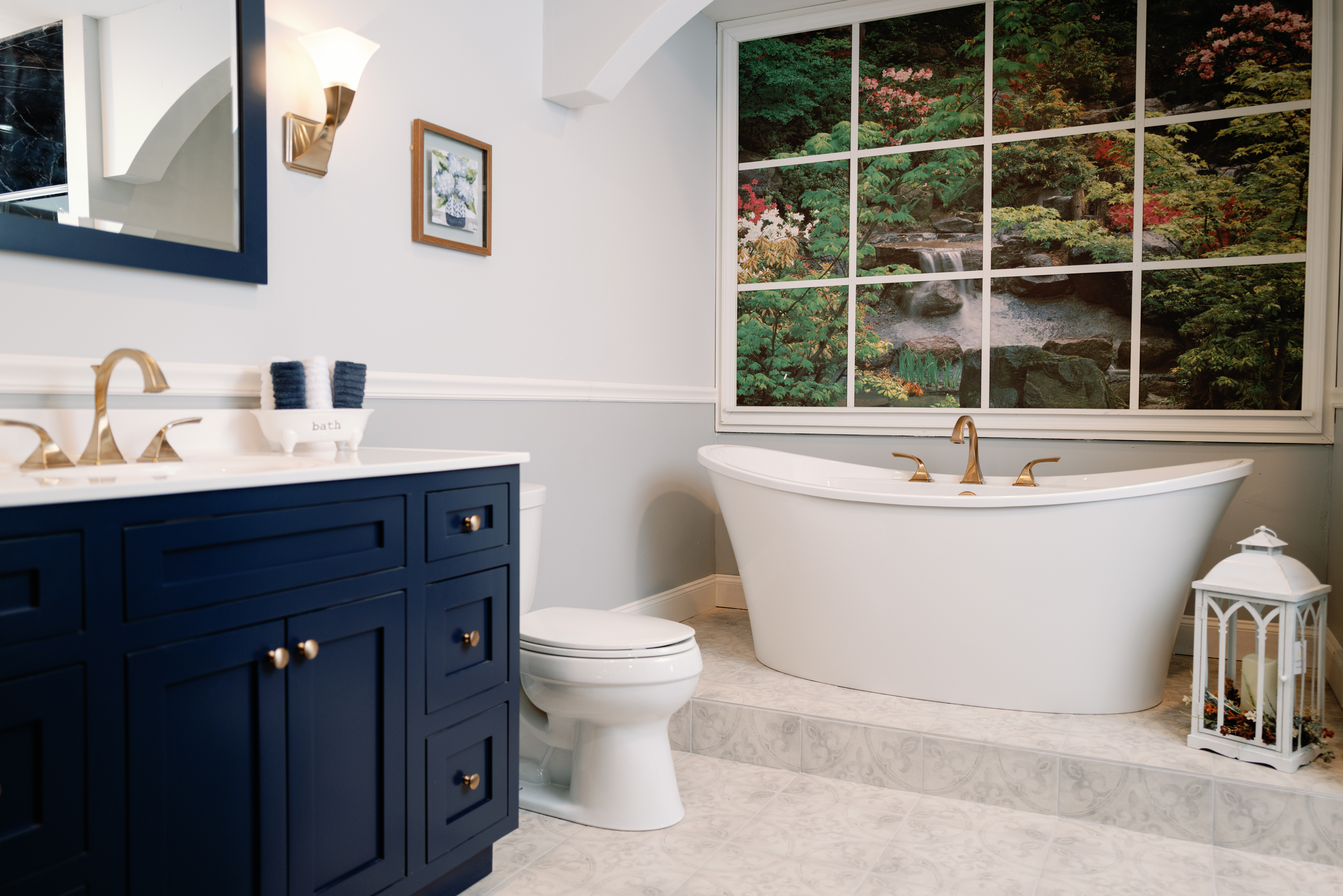 Bath showroom display featuring a freestanding soaking tub, navy vanity with brushed gold faucets, and decorative window scene